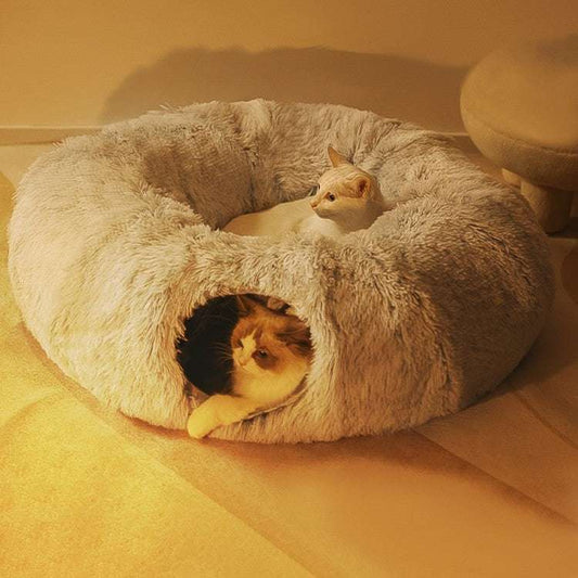 Two cats inside a fluffy donut-shaped pet bed on a soft surface.